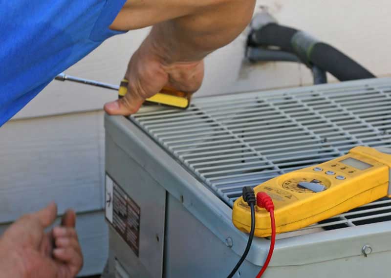 HVAC technician performing AC maintenance on an outdoor condenser using a screwdriver and digital multimeter